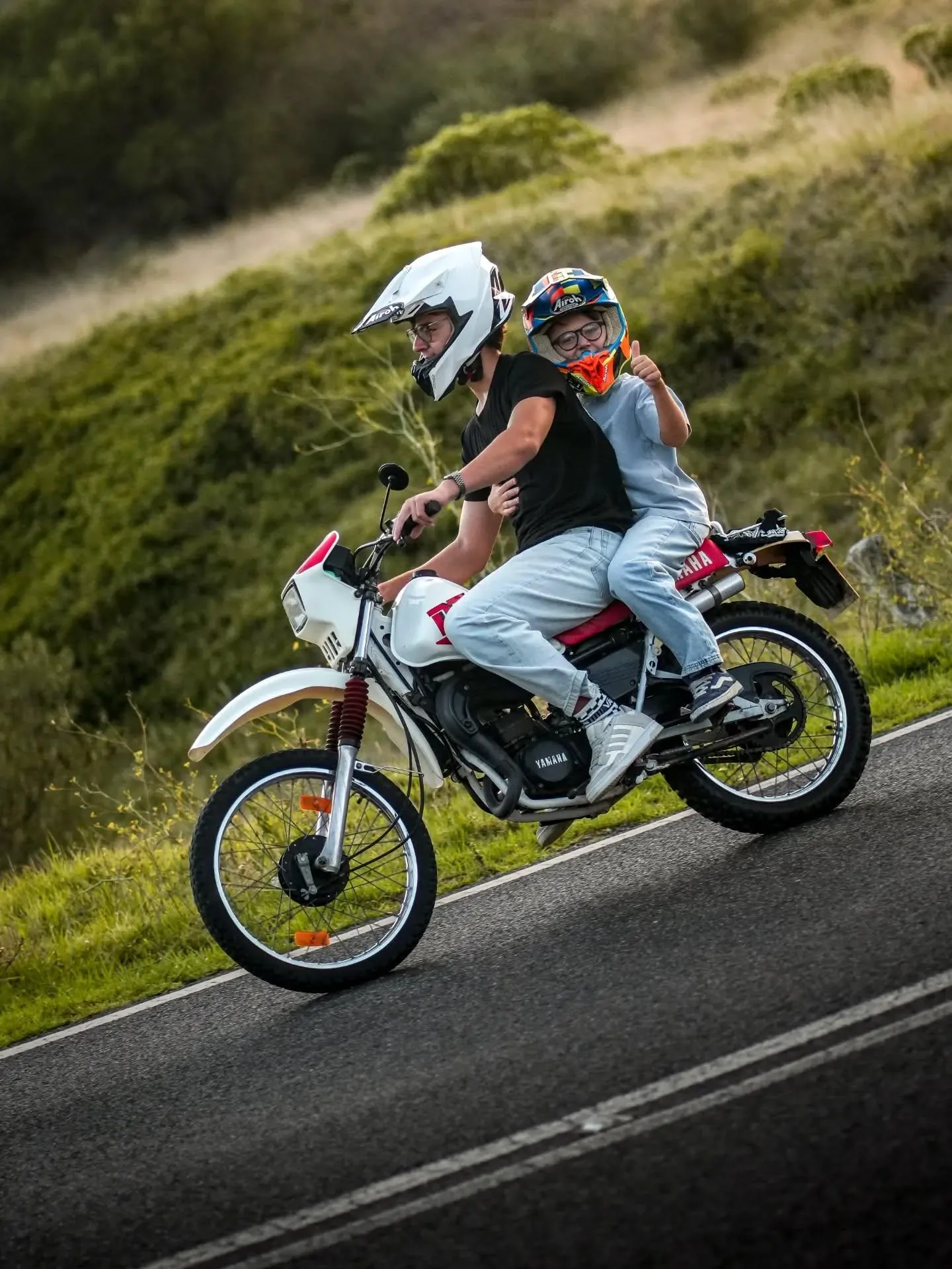 Dois motociclistas de capacete Airoh numa Yamaha clássica branca e vermelha, com o passageiro a fazer sinal de polegar para cima