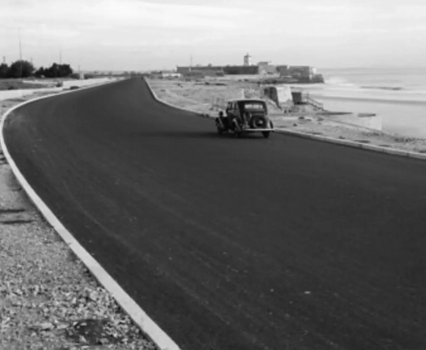 Fotografia antiga da Marginal com asfalto novo, um carro de época e um forte junto à praia