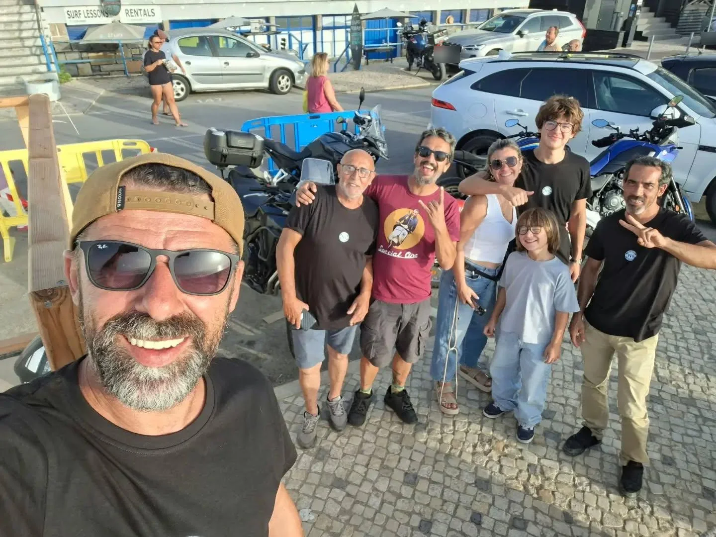 Selfie de grupo de várias gerações do Marginais Motor Club, com t-shirts do clube, junto às motas estacionadas à beira-mar