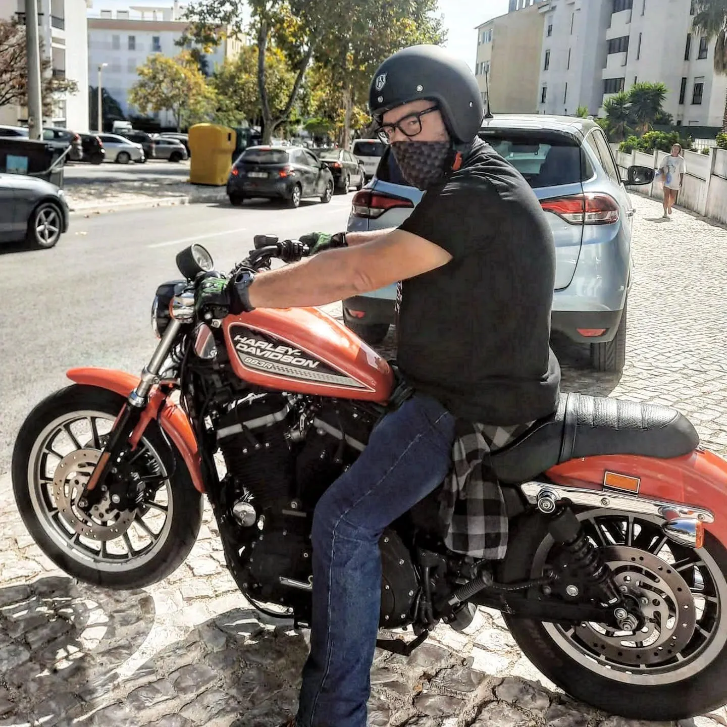 Motociclista de capacete e máscara numa Harley-Davidson Sportster laranja, estacionada numa rua com calçada